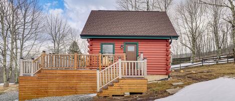 Une cabane en rondins rouge, dotée d’une terrasse en bois et d’une porte verte, entourée d’arbres et de neige.