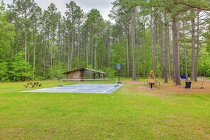 Una cancha de tenis con una cabaña de madera, una mesa de picnic y un conjunto de columpios en un área boscosa.