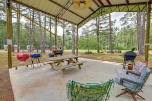 An outdoor covered area with picnic tables, a grill, a chair, and a view of a forest.