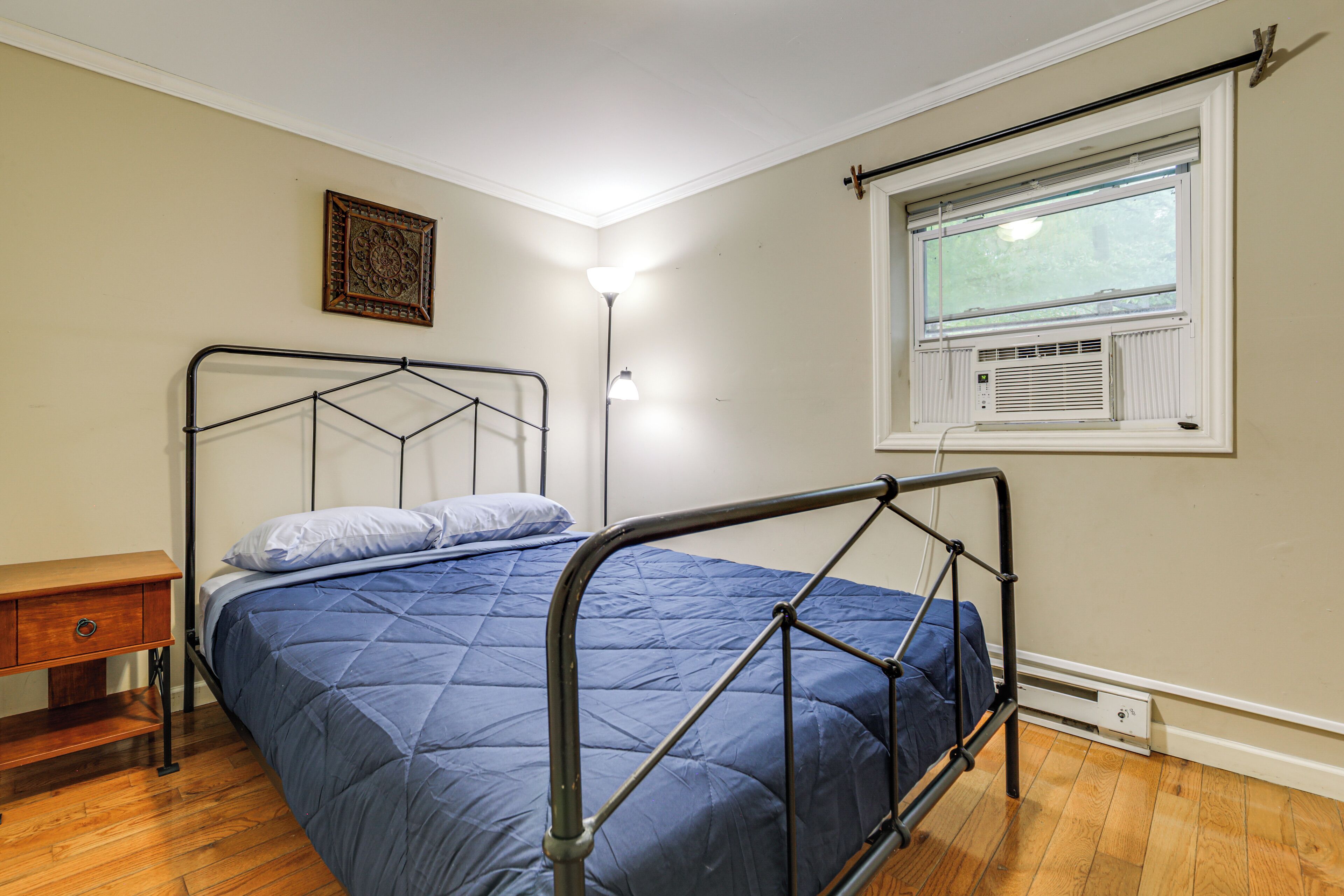 Une chambre à coucher avec un cadre de lit en métal, une table de chevet en bois, une lampe de chevet et une fenêtre équipée d’un appareil de climatisation.