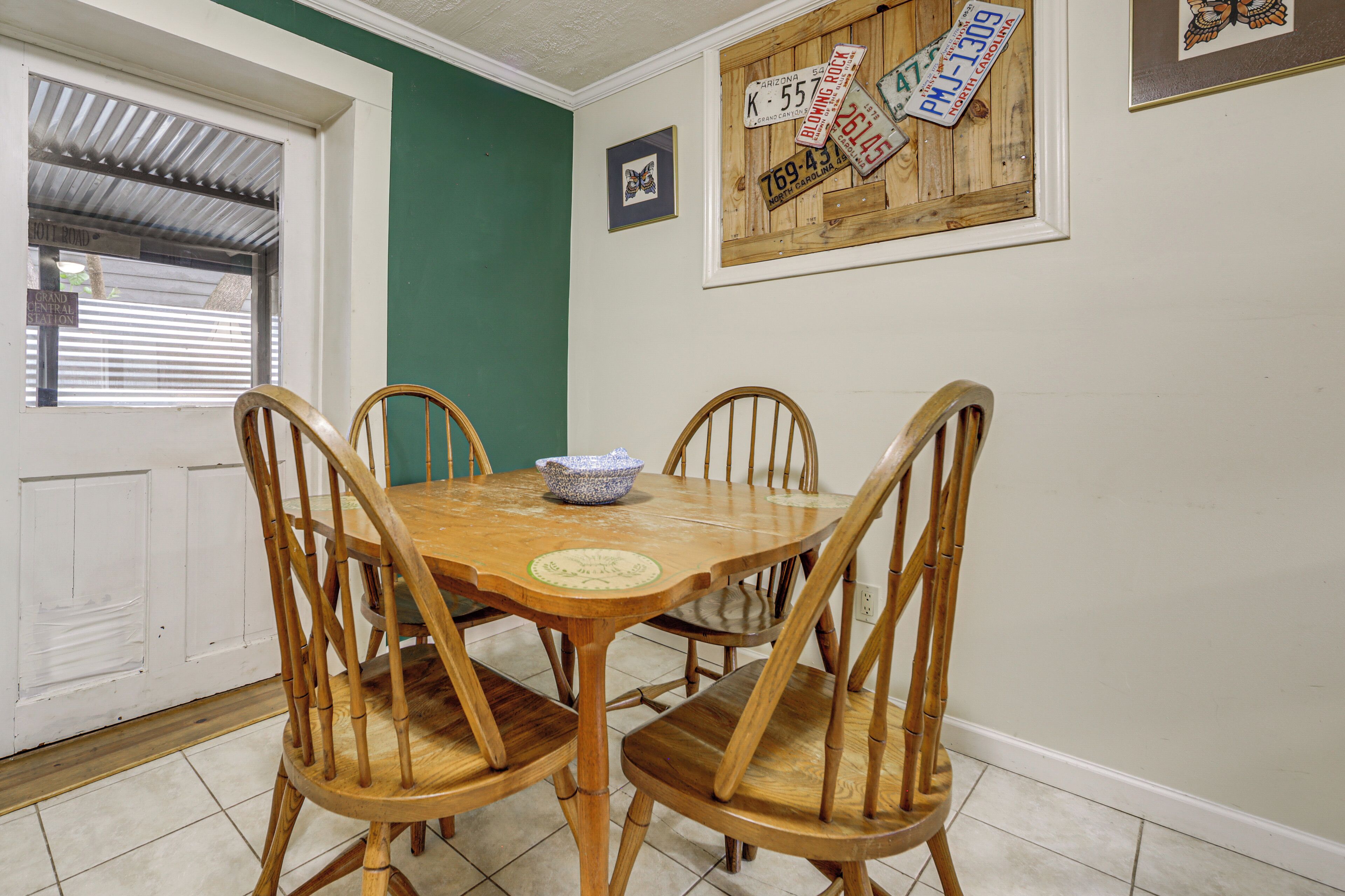 A dining area with a wooden table and chairs, a window with blinds, and a wall with a wooden sign and a framed picture.