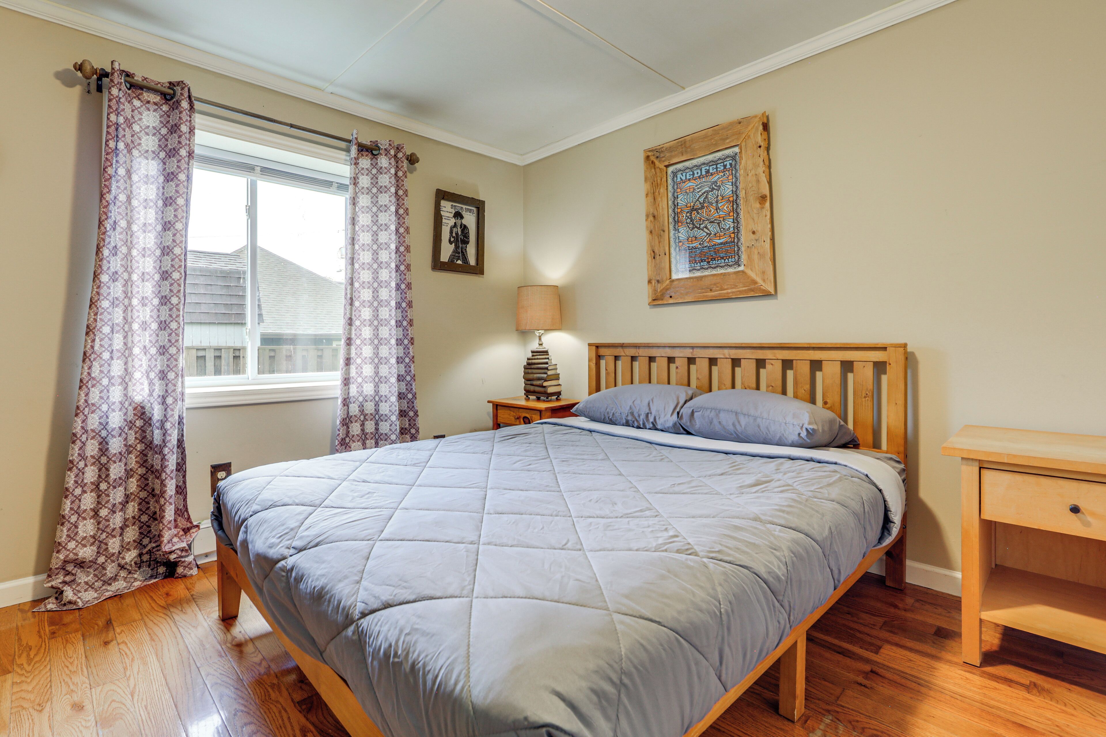 A bedroom with a bed, wooden headboard, a nightstand with a lamp, a framed picture on the wall, and a window with curtains.