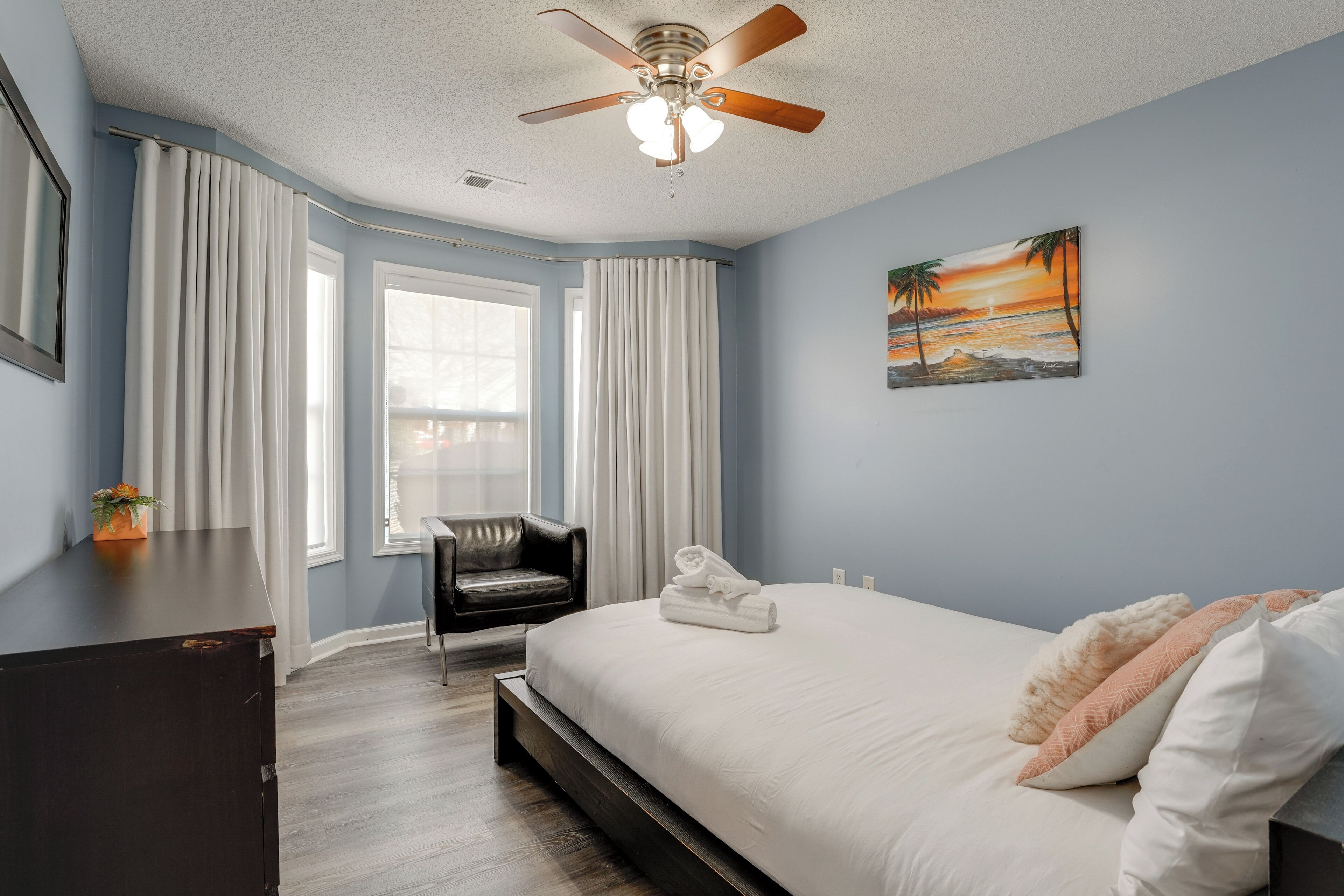 A bedroom with a bed, a ceiling fan, a window with curtains, a chair, and a painting on the wall.