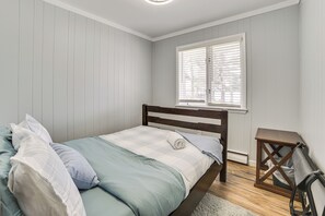 A bedroom with a bed, a window with blinds, a wooden nightstand, and a white rug. - Spacious Getaway w/ Beach & Views on Otsego Lake! (Gaylord)