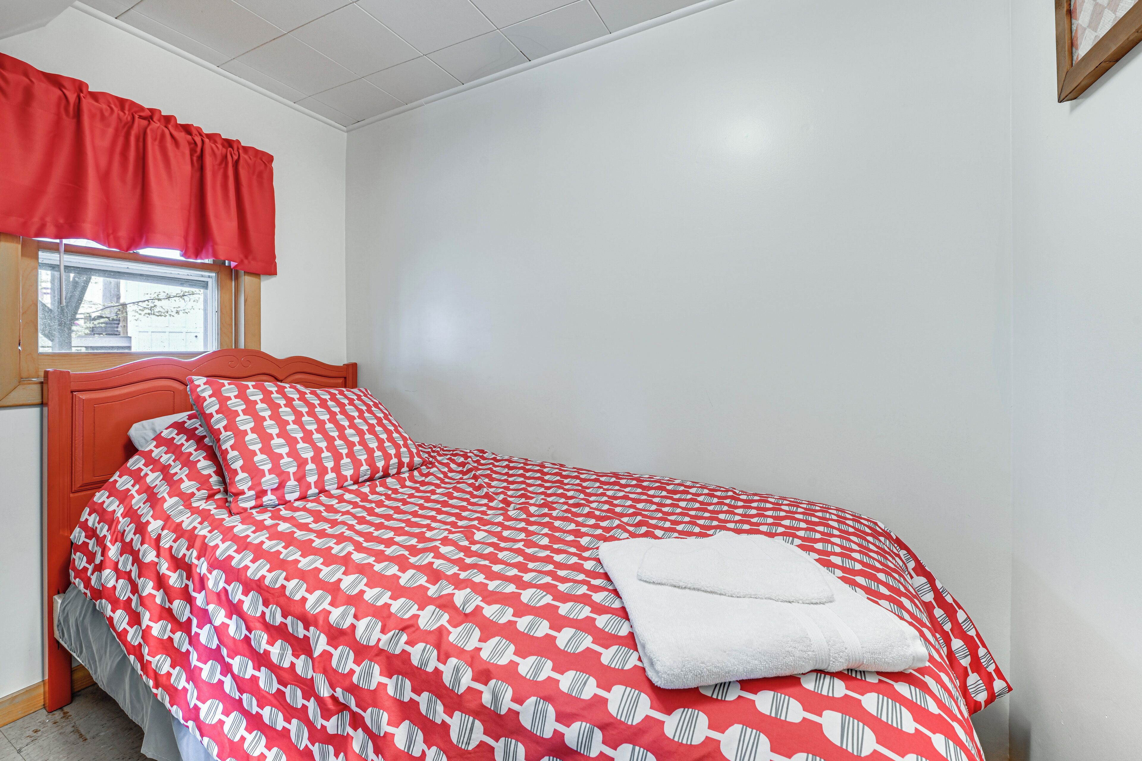Ein ordentlich bezogenes Bett mit einer gemusterten Tagesdecke, ein weißes Handtuch und ein Fenster mit rotem Vorhang.