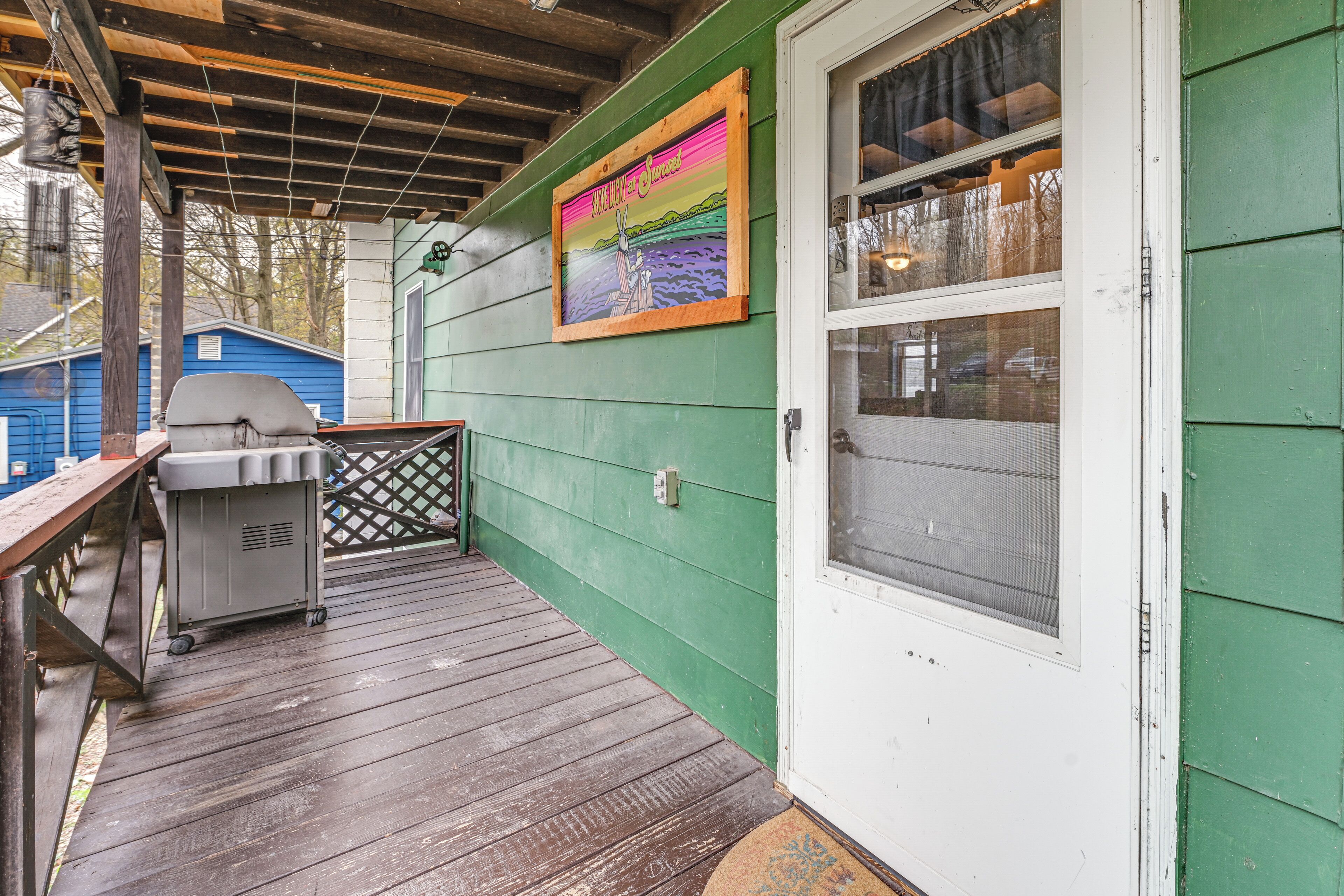 Una terraza de madera con una parrilla, una puerta con ventanal y un letrero colorido en la pared.