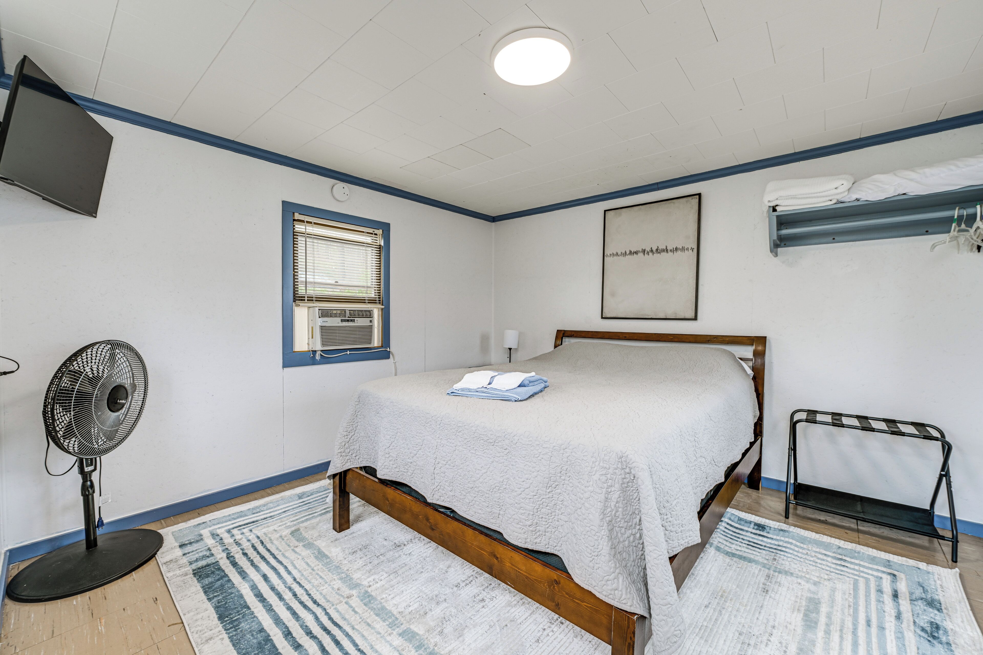 A bedroom with a bed, a standing fan, a window, a wall-mounted TV, and a bunk bed.