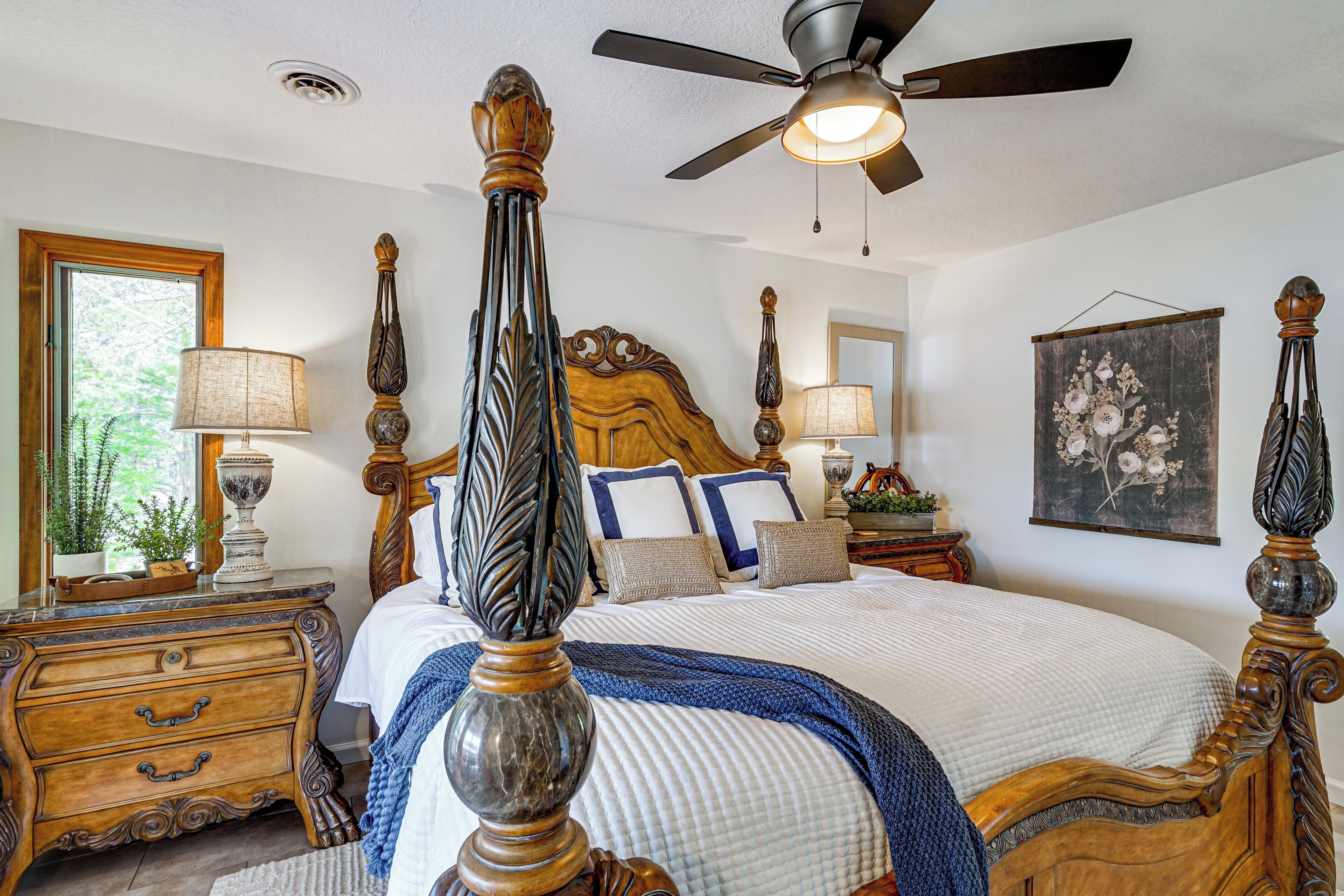 A bedroom with a four-poster bed, a ceiling fan, a large window, and a framed floral artwork on the wall.