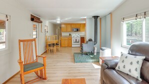A spacious living area with a wooden rocking chair, a leather sofa with a pillow, a kitchen with wooden cabinets, and a large window offering a view. - Deschutes Forest Basecamp! Oregon Cabin (La Pine)