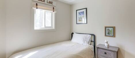 A single bed with a white comforter, a nightstand with drawers, a window with a name sign, and a framed picture on the wall.