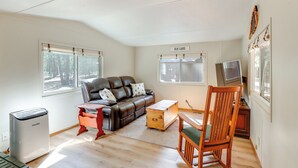 A room with a leather sofa, a wooden rocking chair, a small wooden table, a television, and a window with a view of trees. - Deschutes Forest Basecamp! Oregon Cabin (La Pine)