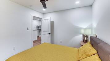 Un dormitori amb un llit, un ventilador de sostre, una tauleta de nit amb una làmpada i una porta que dona a una altra habitació.