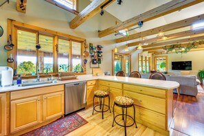 A spacious kitchen with wooden cabinets, a large island with stools, a dishwasher, and a view of the outdoors through large windows.