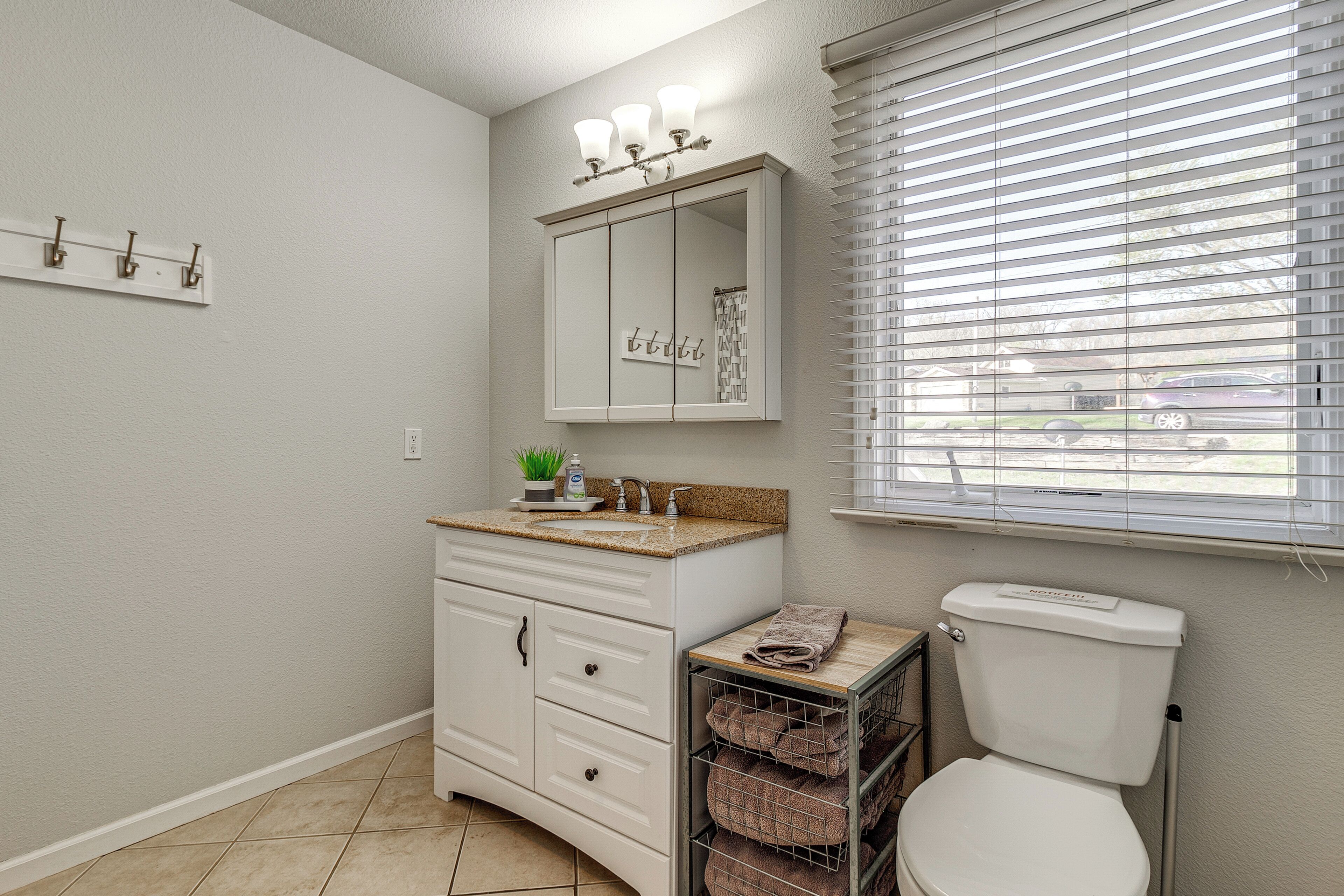 Une salle de bain avec un meuble-lavabo blanc, un miroir avec éclairage intégré, des toilettes et une fenêtre avec des stores.