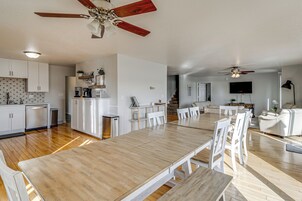 Amplia sala de estar con mesa de comedor de madera, sillas blancas, ventilador de techo y área de cocina con gabinetes blancos y lavavajillas.