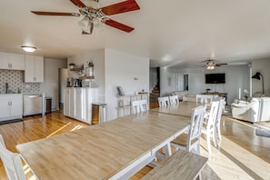 A spacious living area with a wooden dining table, white chairs, a ceiling fan, and a kitchen area with white cabinets and a dishwasher.