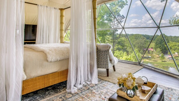 Quarto com cama, televisão, cadeira e vista para o verde através de uma grande janela.