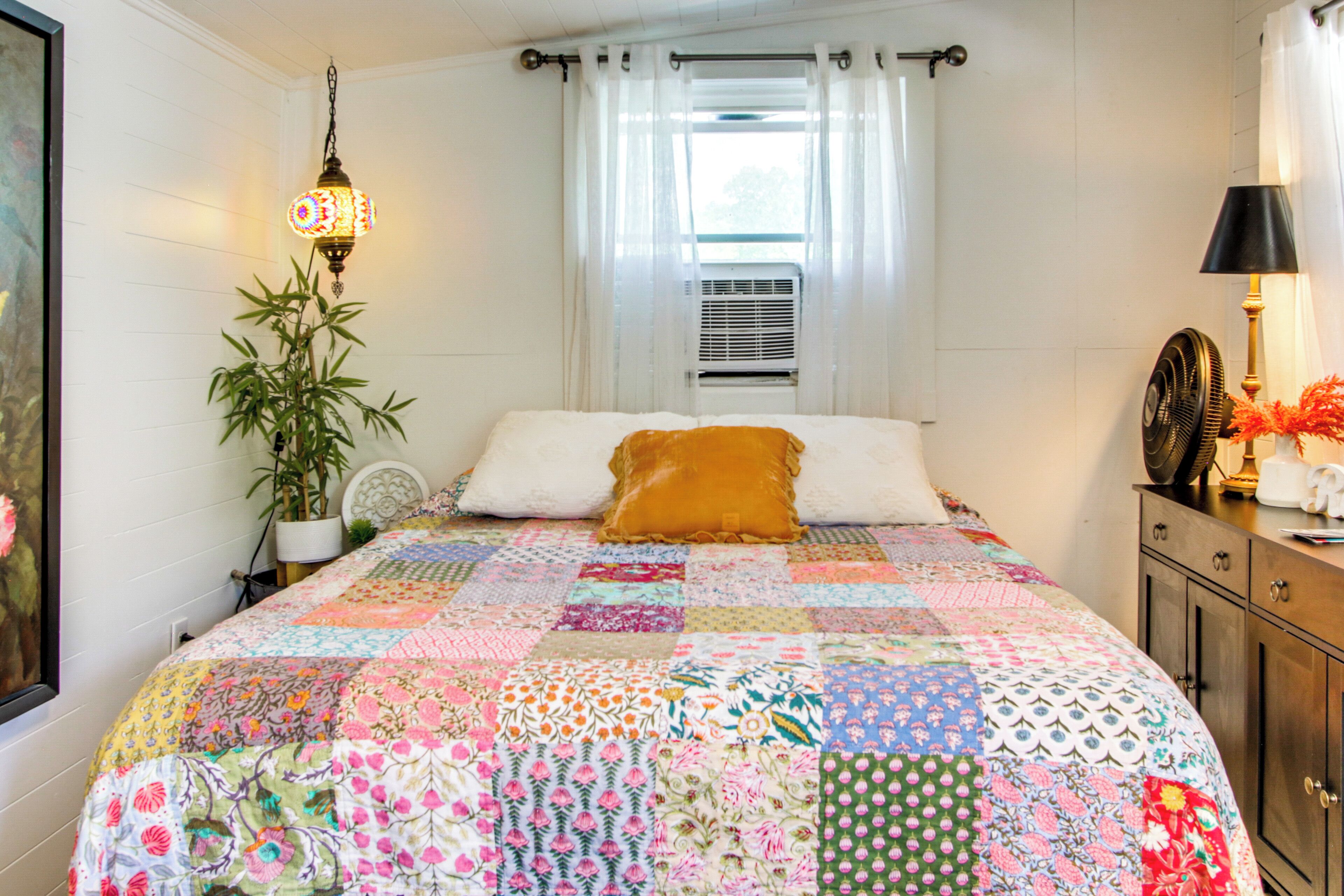 A bedroom with a quilted bedspread, a potted plant, a wall-mounted lamp, and a bedside table with a lamp.