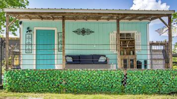 A small, rustic building with a teal door, a wooden bench with cushions, and a green leafy wall in front.