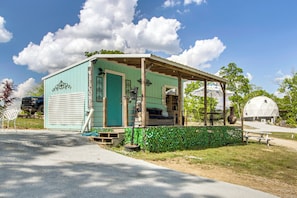 A small, single-story structure with a porch and a covered seating area, surrounded by greenery and a clear sky.