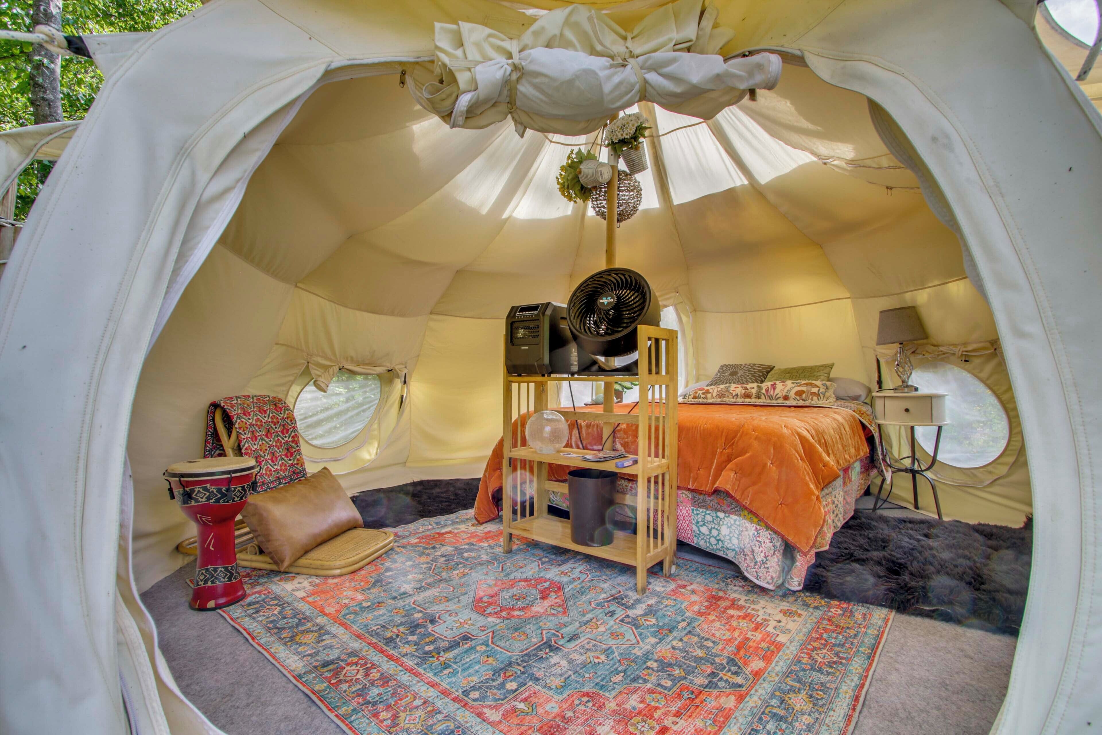 A tent with a bed, a small table, a chair, a fan, and a rug.