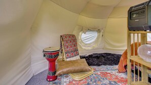 Ein Zeltinnenraum mit einem gemusterten Teppich, einer roten Trommel, einer gepolsterten Sitzgelegenheit, einem kleinen Regal mit einem dekorativen Gegenstand und einem Fenster.