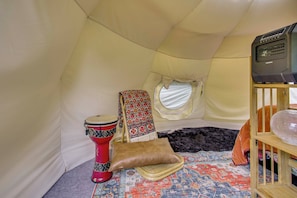 A tent interior with a patterned rug, a red drum, a cushioned seat, a small shelf with a decorative item, and a window.