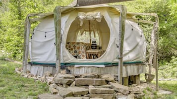 A rustic tent with a table and chairs inside, surrounded by rocks and greenery.