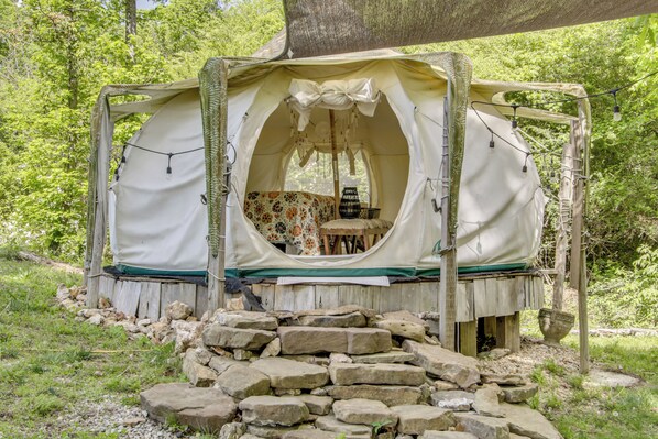 A rustic tent with a table and chairs inside, surrounded by rocks and greenery.
