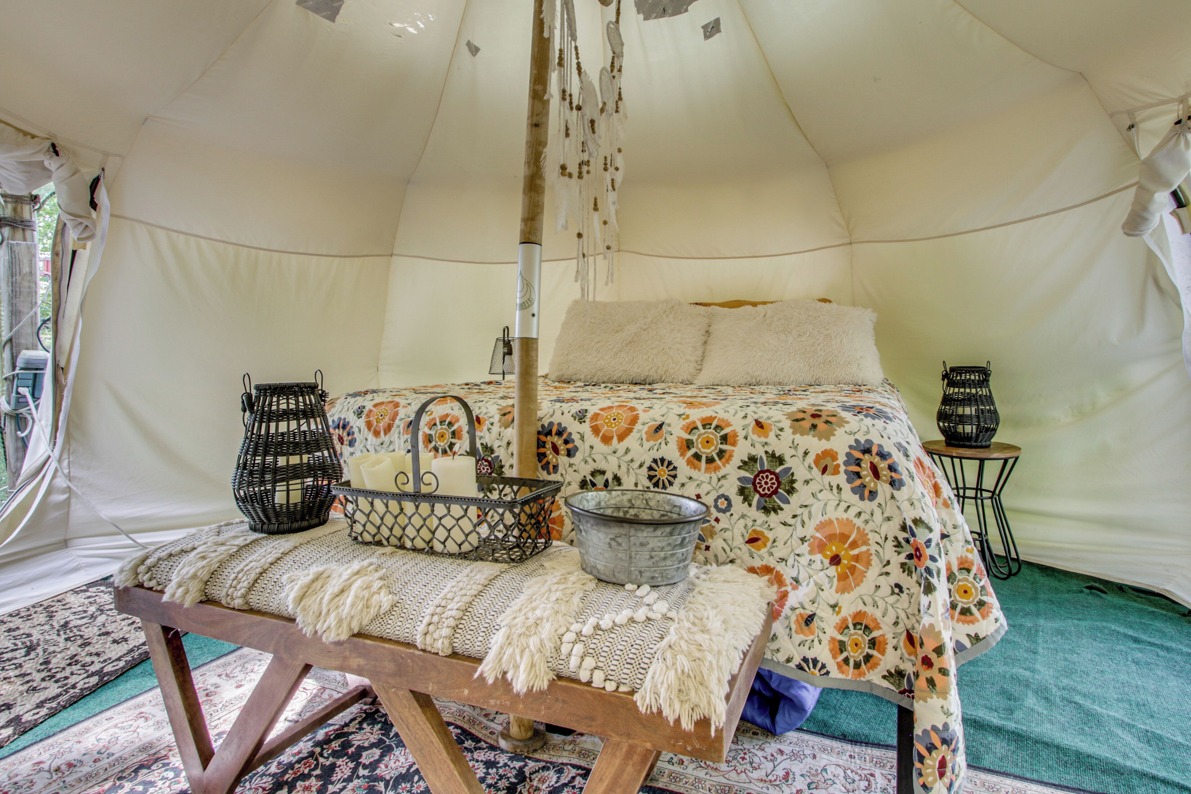 A tent with a bed, a bench, a hanging light, and a small table with a basket and candle.