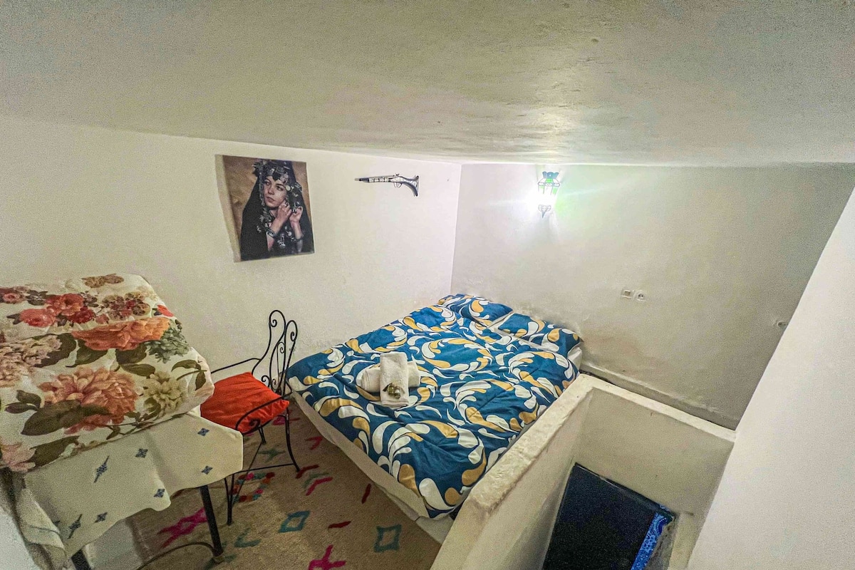 A small, compact room with a bed, a desk, and a chair. There is a small window with a view of the outside.