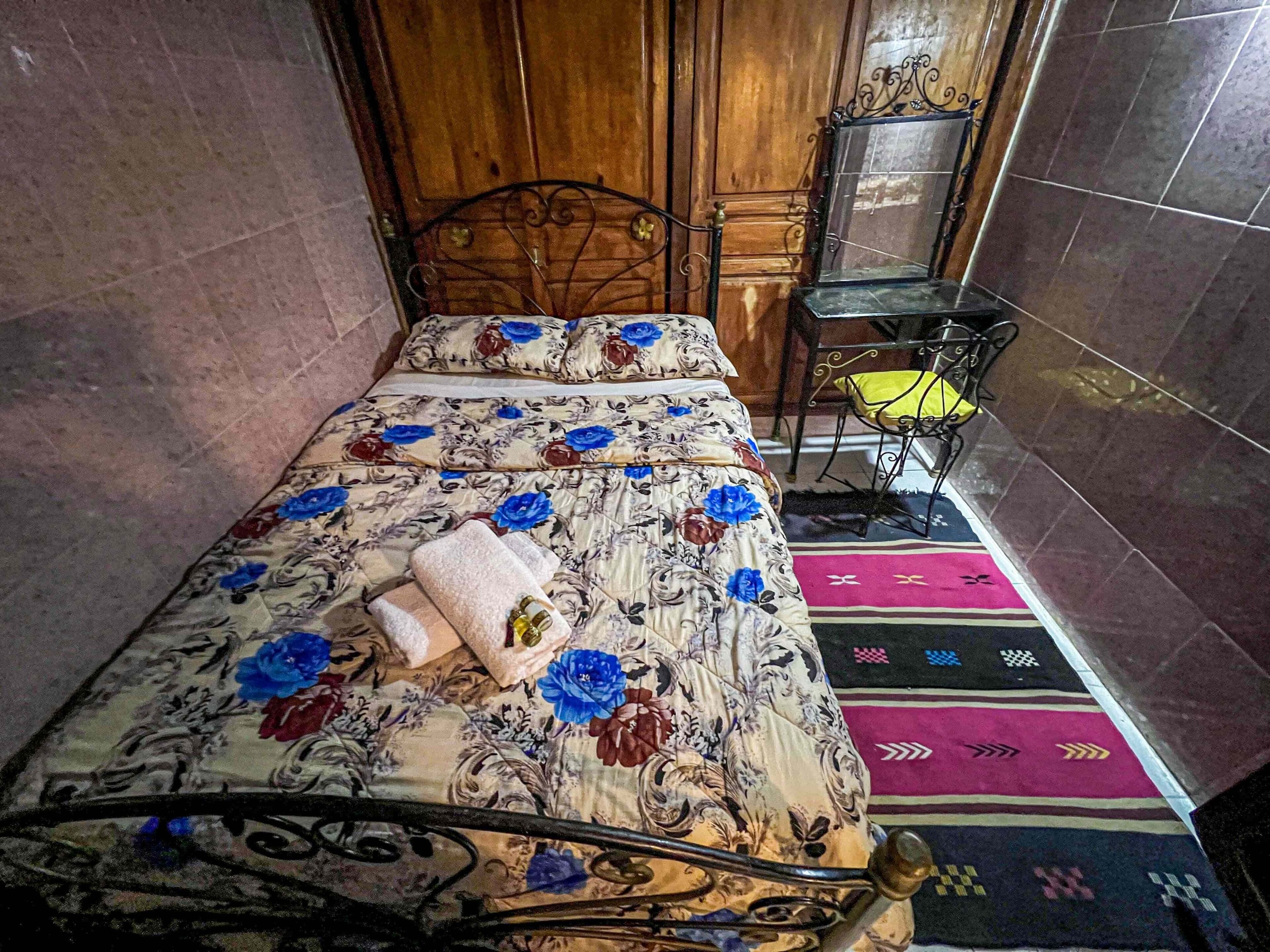 Uma cama de solteiro com colcha florida, cabeceira de madeira, uma mesinha com almofada amarela e um tapete estampado.