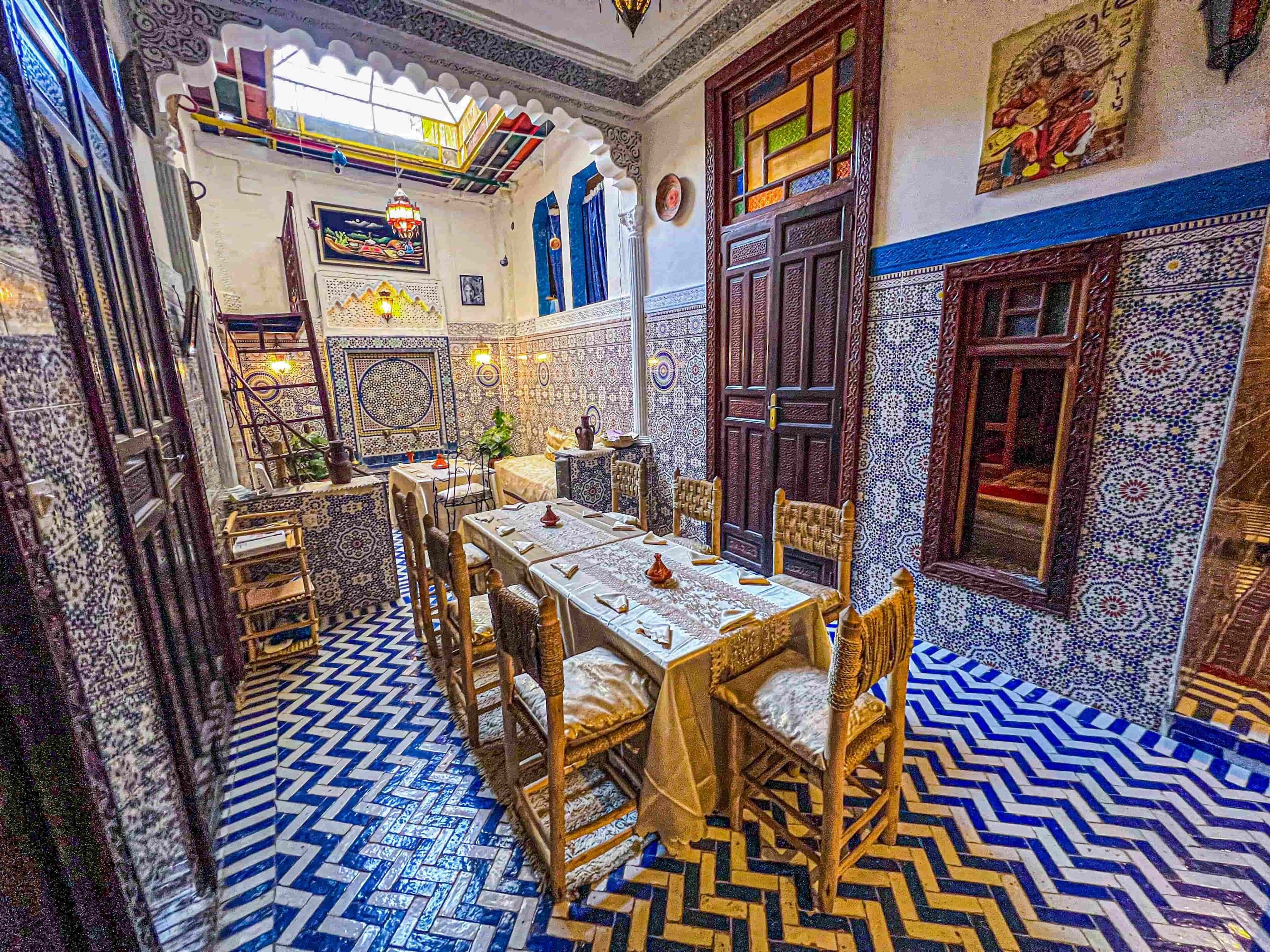Um interior tradicional marroquino com soalho em azulejos, zona de refeições com mesa e cadeiras, e decoração de parede elaborada.