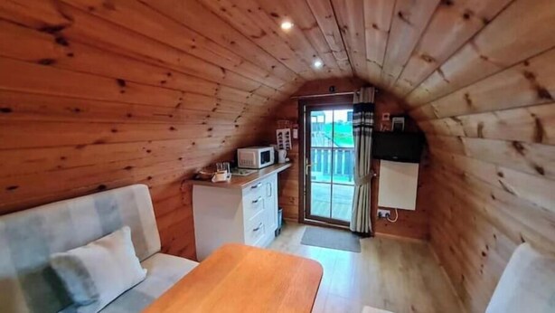 A cozy cabin with a wooden interior, a sofa, a wooden table, a kitchenette, and a sliding glass door leading outside.