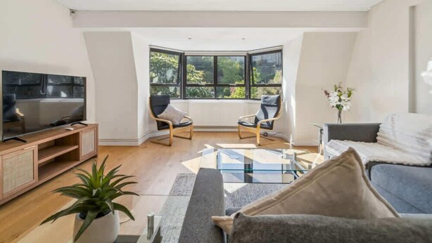 A modern living room with a television, two armchairs, a sofa, a glass coffee table, and a large window offering a view of greenery. - Windsor Castle Views - Spacious Flat - By Tempstay (Windsor)