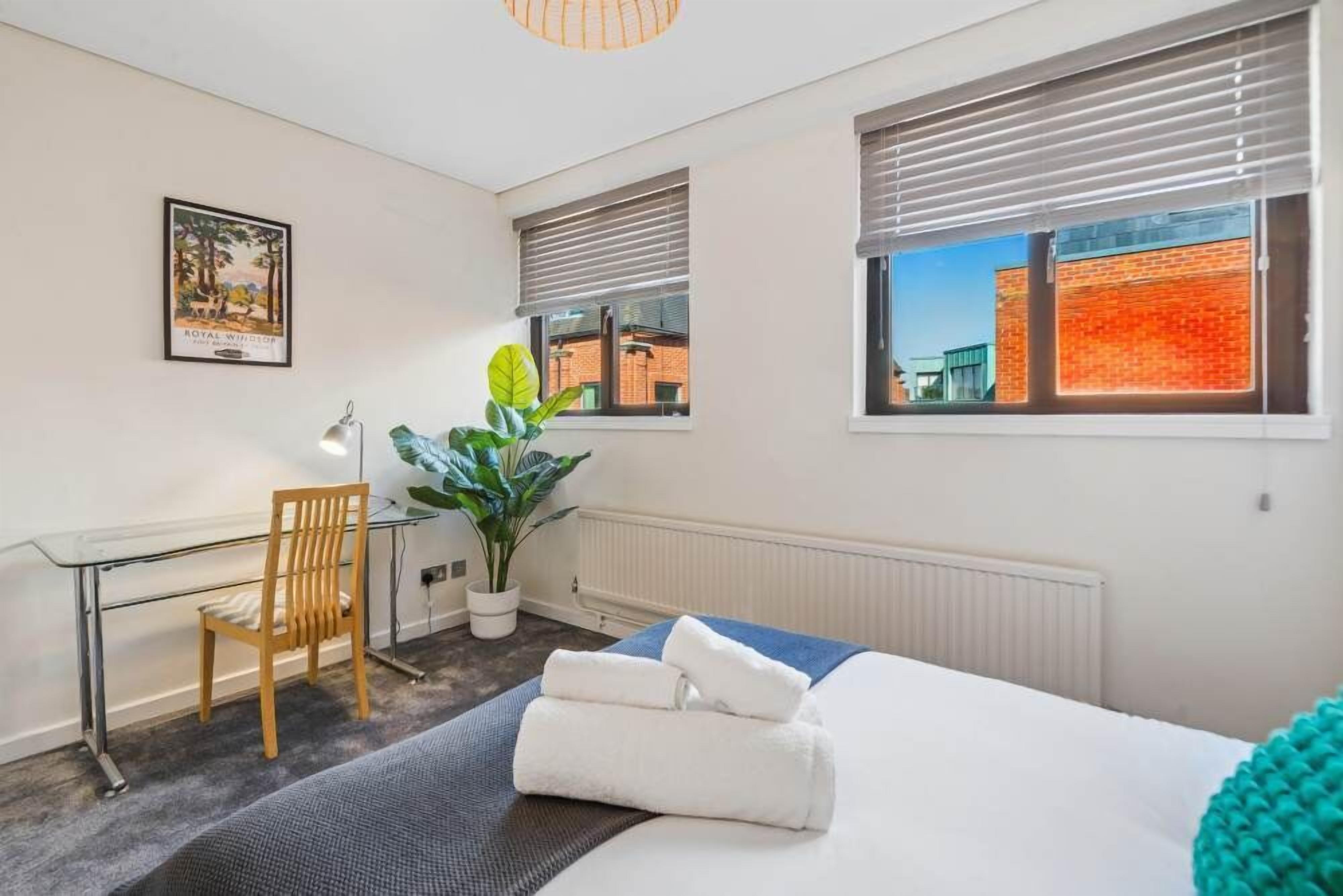 Une chambre d’hôtel avec un lit, un bureau, une chaise, une plante en pot et une fenêtre avec des stores.