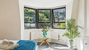 A bedroom with a bed, a radiator, a window with a view of greenery, a small table with a vase, and a potted plant. - Windsor Castle Views - Spacious Flat - By Tempstay (Windsor)
