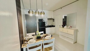 Una cocina moderna con armarios blancos, un refrigerador negro y un microondas. Hay un comedor con una mesa de madera preparada para cuatro personas, sillas blancas y lámparas colgantes.