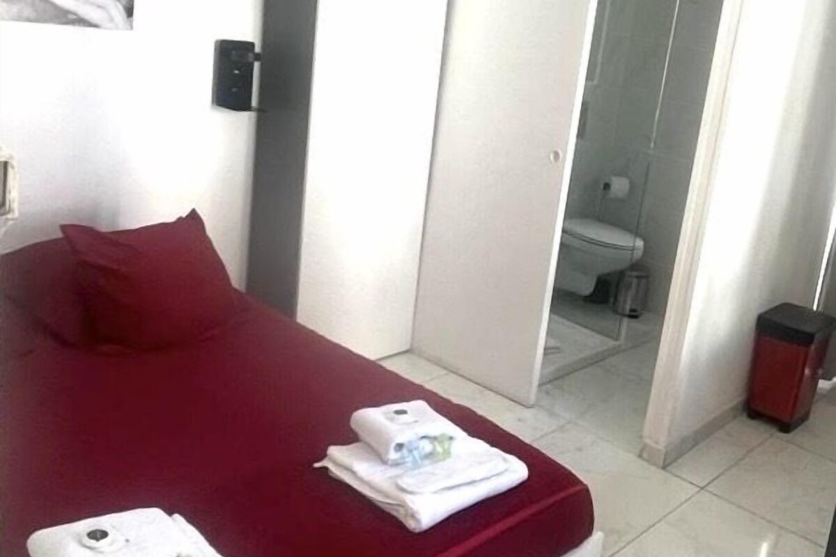 Habitación de hotel con cama, cabecera roja, pared blanca con un cuadro enmarcado y un baño visible a través de una puerta abierta.