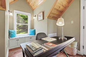 Ein Zimmer mit Schreibtisch, Fensterbank, Lampe und einem gerahmten Bild an der Wand.