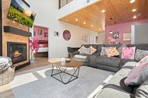 A modern living room with a fireplace, a sofa set, a coffee table, and a large window overlooking a garden. - Luxury Dolly-Themed Cabin, Indoor Pool, Hot Tub (Sevierville)