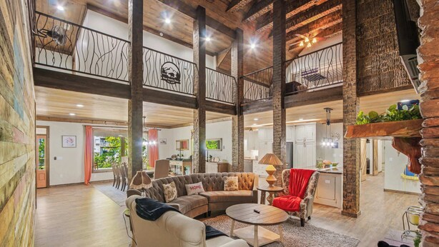 A spacious loft-style living area with a high ceiling, exposed wooden beams, a large sectional sofa, a round coffee table, and a red armchair. - Luxe Riverfront Cabin ・ Walk to Parkway・Hot Tub (Pigeon Forge)