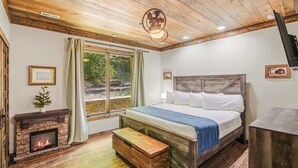 A bedroom with a bed, a fireplace, a window with curtains, and a ceiling fan. - Luxe Riverfront Cabin ・ Walk to Parkway・Hot Tub (Pigeon Forge)