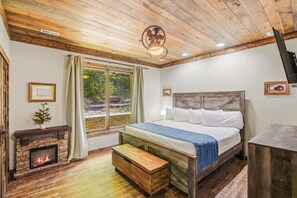 A bedroom with a bed, a fireplace, a window with curtains, and a ceiling fan. - Luxe Riverfront Cabin ・ Walk to Parkway・Hot Tub (Pigeon Forge)