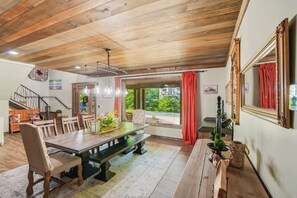 Un comedor con mesa y sillas de madera, un banco, una araña y vistas a través del ventanal.