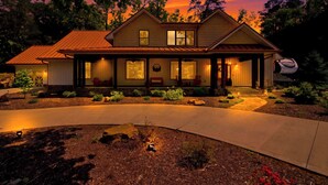 A two-story house with a covered porch, large windows, and a well-lit driveway. - Luxe Riverfront Cabin ・ Walk to Parkway・Hot Tub (Pigeon Forge)