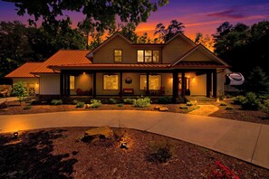 A two-story house with a covered porch, large windows, and a well-lit driveway. - Luxe Riverfront Cabin ・ Walk to Parkway・Hot Tub (Pigeon Forge)