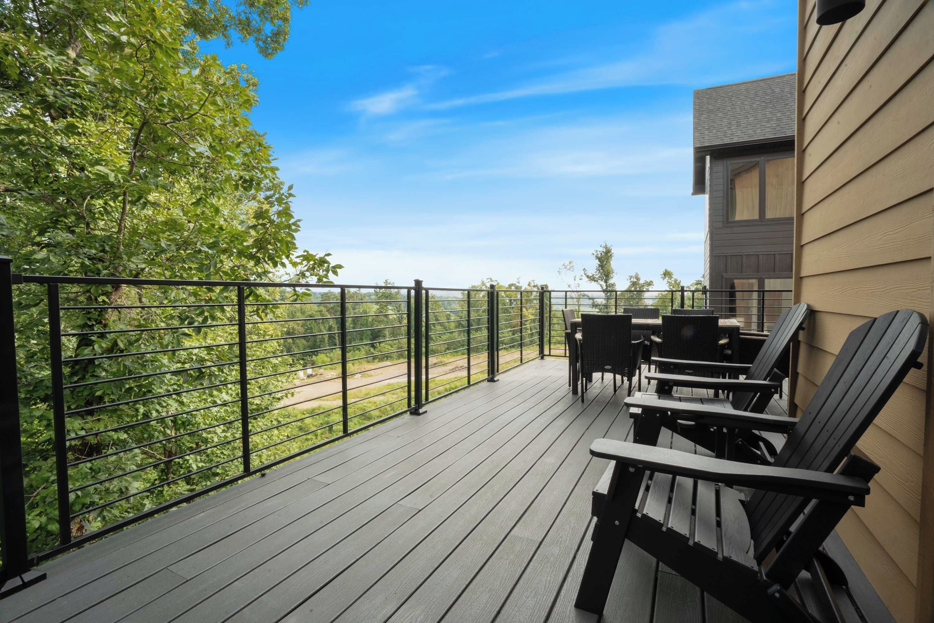 Terraza de madera con mobiliario exterior, baranda metálica y vista a árboles y un paisaje distante.