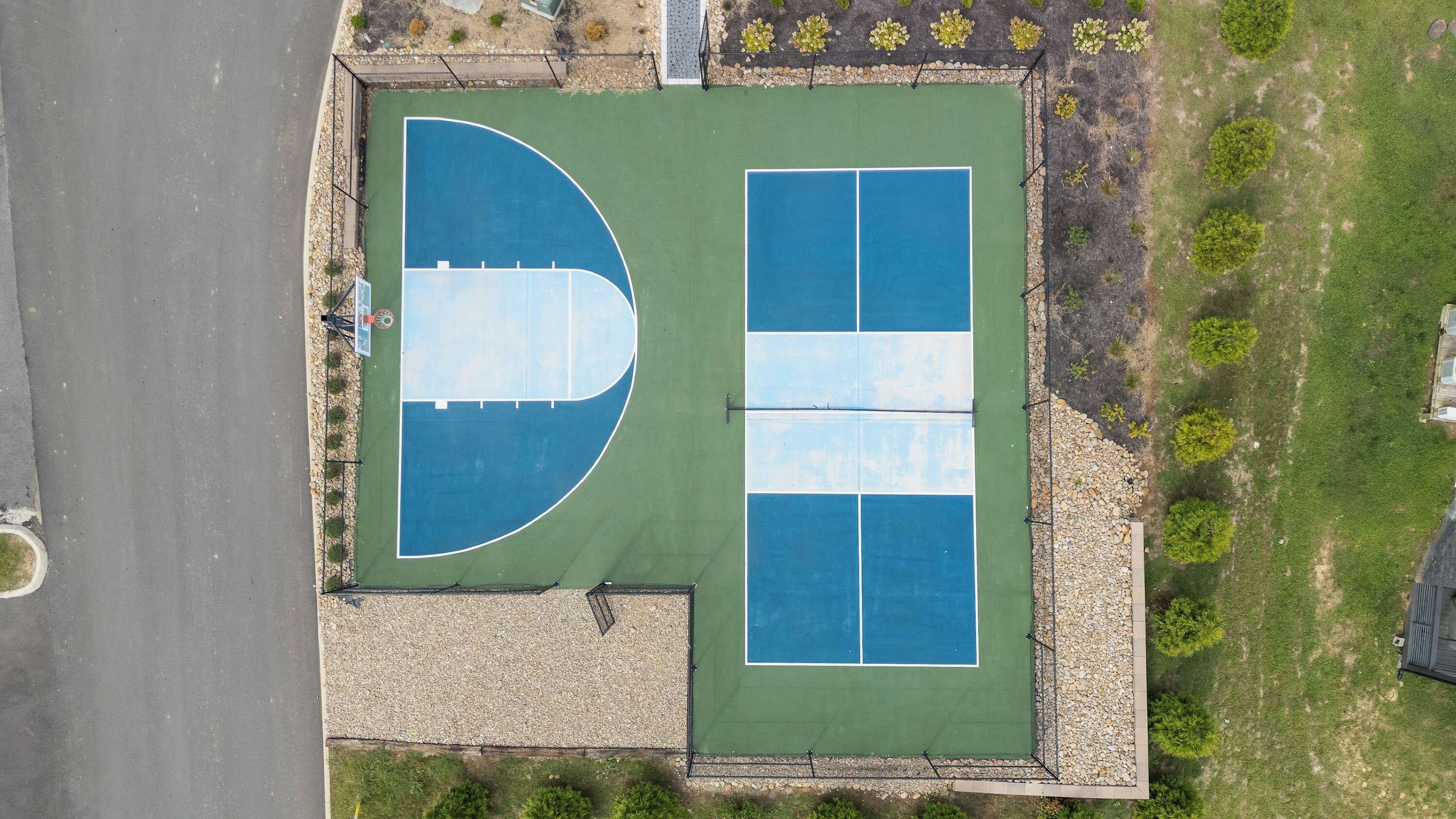 An aerial view of a sports court with a basketball court and a tennis court, surrounded by a gravel path and greenery.