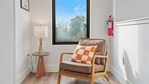 A corner room with a wooden chair, a small table with a lamp, a window with a view of trees, and a fire extinguisher on the wall.